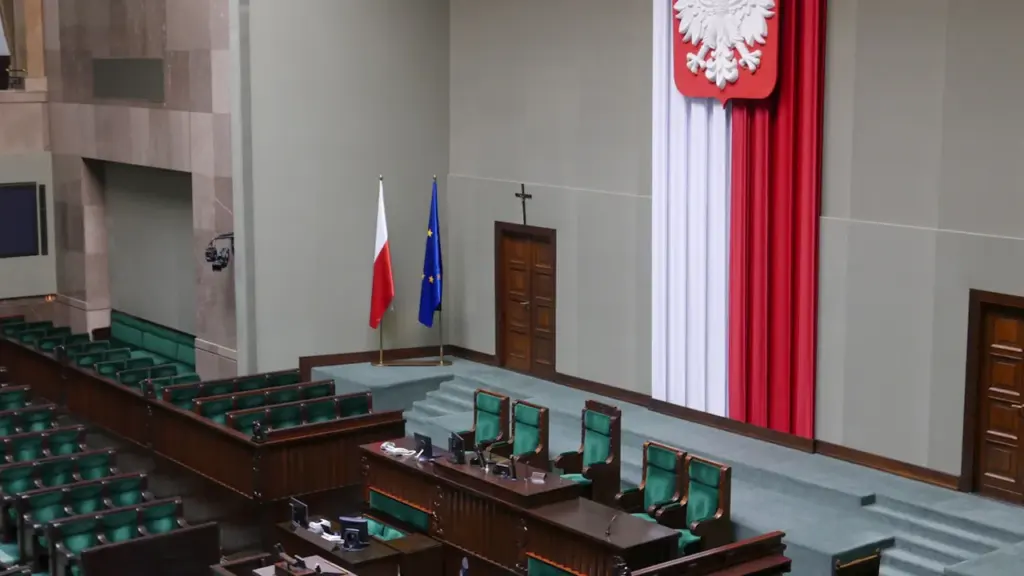 Sala plenarna polskiego Sejmu z biało-czerwoną flagą i godłem Polski na ścianie.